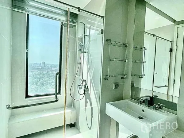 Condominium for rent in Bangkok - Modern bathroom with glass shower enclosure, large window and city panorama.