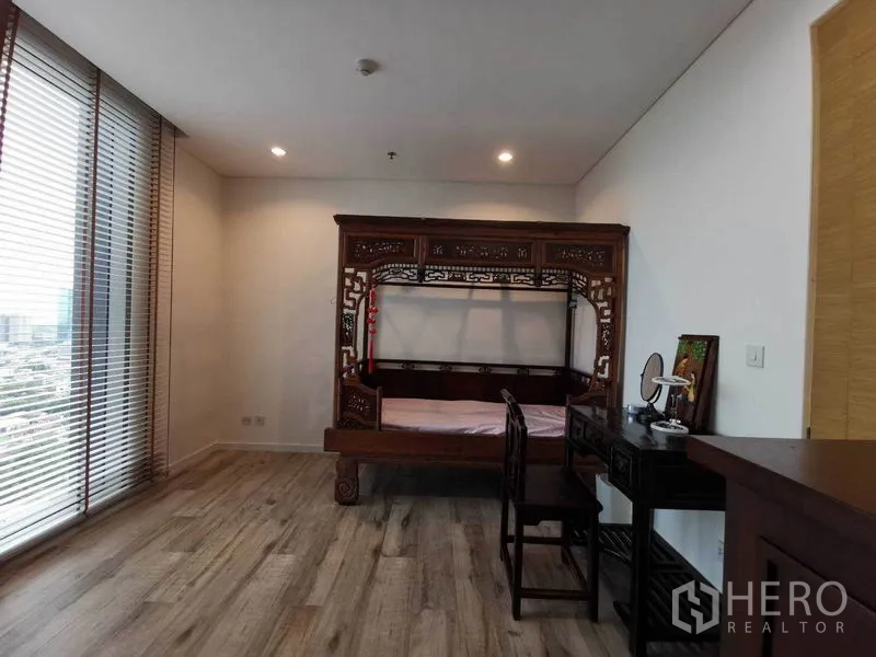 Condominium for rent or sale in Bang Rak, Bangkok - Bright bedroom with floor-to-ceiling windows, wood flooring, and an ornate carved wooden bed with desk.