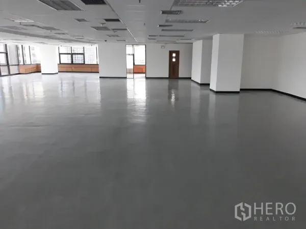 Office Space for rent in Bangkok - Large empty office space with multiple pillars, drop ceiling, and wide windows bringing in natural light and a doorway to a service room.
