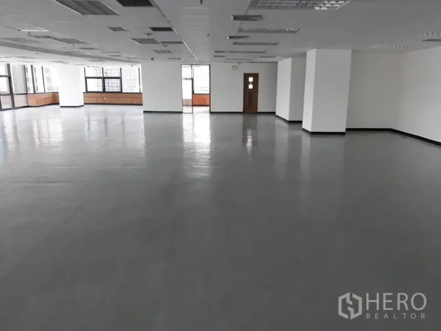 Office Space for rent in Bangkok - Large empty office space with multiple pillars, drop ceiling, and wide windows bringing in natural light and a doorway to a service room.