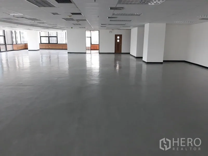 Office Space for rent in Bangkok - Large empty office space with multiple pillars, drop ceiling, and wide windows bringing in natural light and a doorway to a service room.
