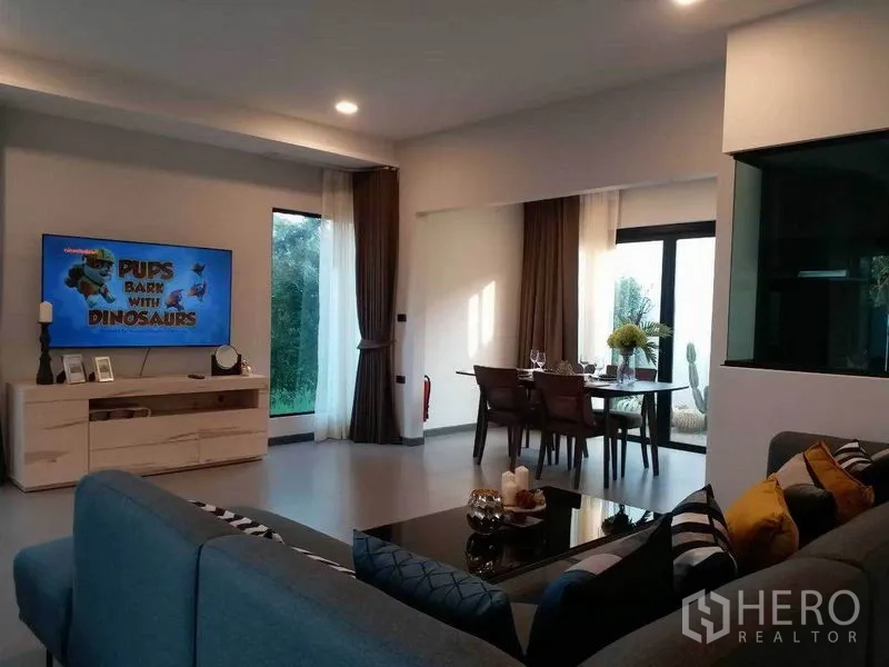 Townhouse for rent in Bang Phli, Samut Prakan - Open-plan living and dining area with sectional sofa, wall-mounted TV, and large windows with curtains.