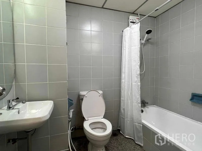 Condominium for rent in Ratchathewi, Bangkok - Bathroom with bathtub and shower, white curtain, toilet, and light gray tiled walls.