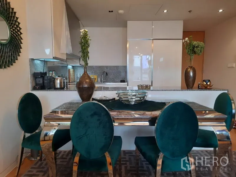 Condominium for rent or sale in Sathon, Bangkok - Dining area with emerald chairs and a polished table beside an open‑plan kitchen with white cabinetry and granite counters.