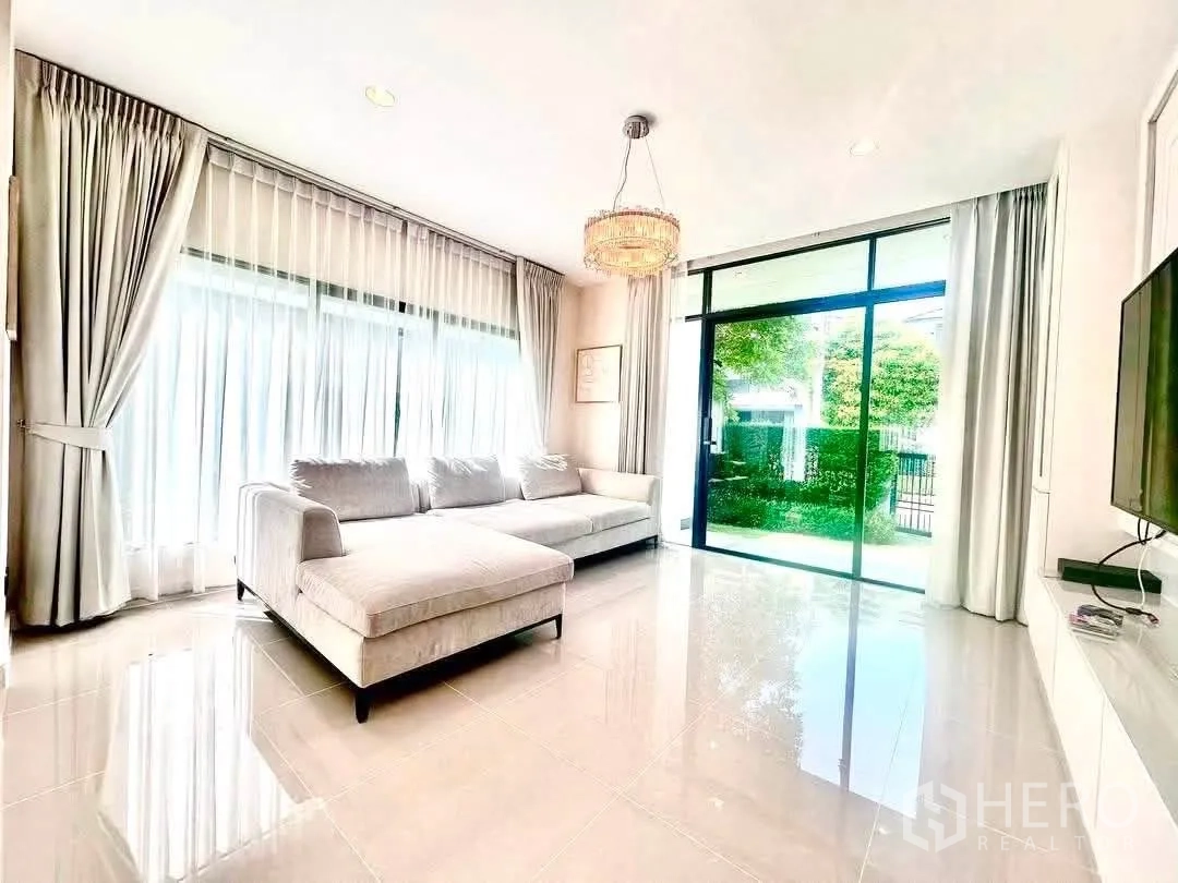 Detached House for rent in Bang Kapi, Bangkok - Living room with L-shaped sofa, sheer curtains and sliding doors to the garden.