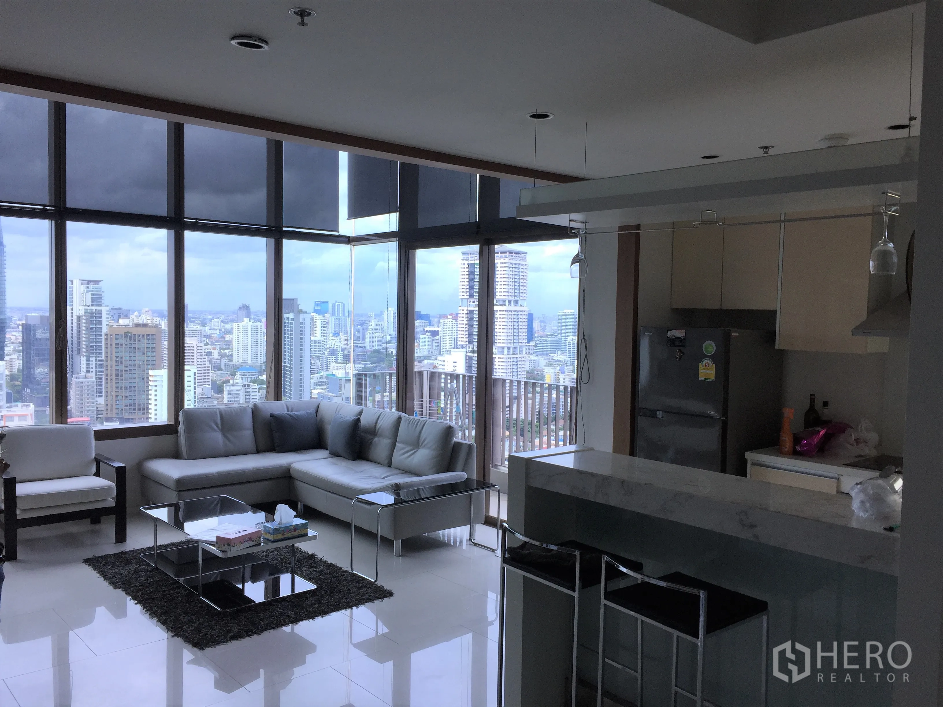 Condominium for rent or sale in Khlong Toei, Bangkok - Open-plan living and kitchen with corner glass walls, L-shaped sofa and panoramic skyline views.