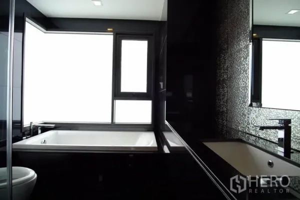 Condominium for rent in Khlong Toei, Bangkok - Modern bathroom with black tiles and a large soaking tub beside a frosted window.