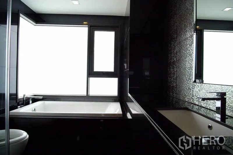 Condominium for rent in Khlong Toei, Bangkok - Modern bathroom with black tiles and a large soaking tub beside a frosted window.