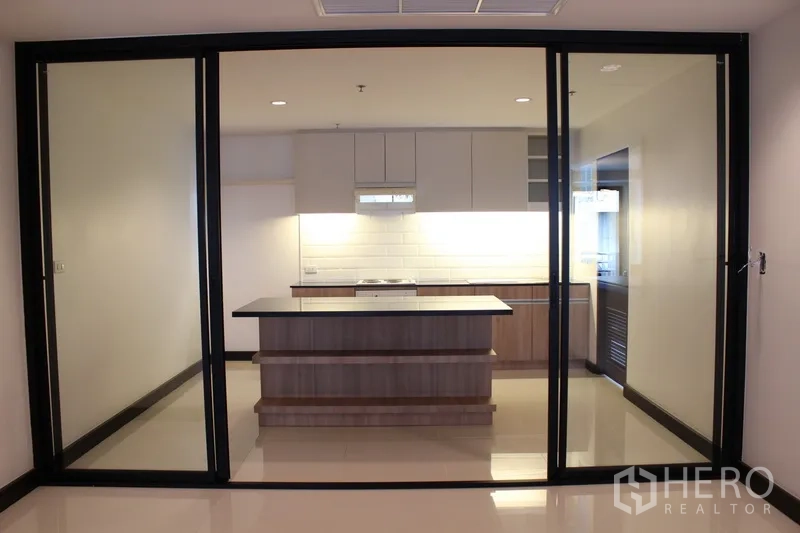 Condominium for rent in Watthana, Bangkok - Enclosed kitchen behind black-framed sliding glass doors with a central island and bright cabinetry.