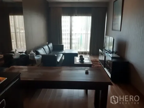 Condominium for rent in Bangkok - Living room with L-shaped leather sofa, TV console, wooden floors and floor-to-ceiling window with sheer curtains.