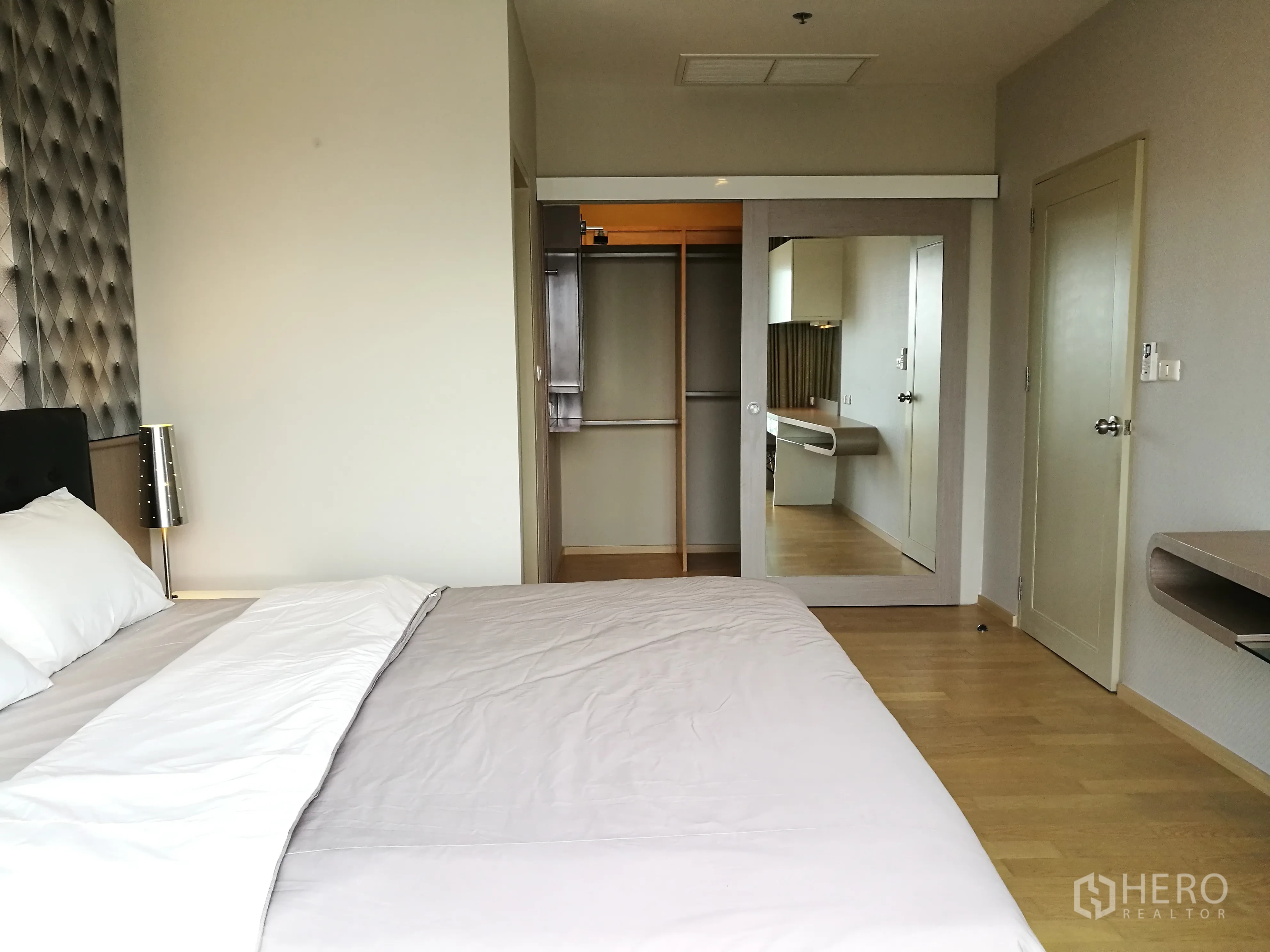 Condominium for rent in Watthana, Bangkok - Bright bedroom with upholstered headboard, bedside lamp, built‑in wardrobe with mirrored sliding door, and light wood flooring.