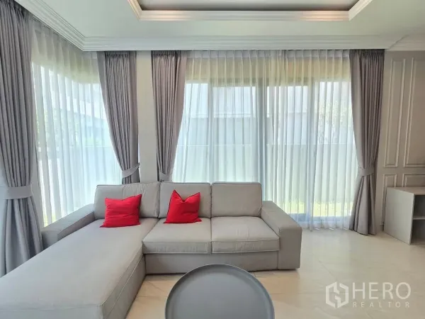 Detached House for rent in Prawet, Bangkok - Close‑up of an L‑shaped gray sofa with red cushions beside tall windows and drapes.