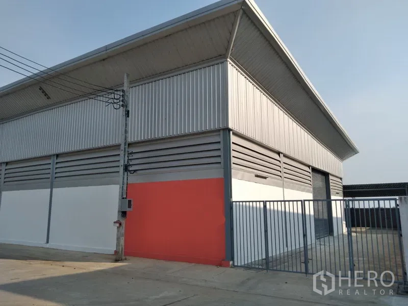 Warehouse for rent in Sai Noi, Nonthaburi - Front exterior of warehouse with orange accent wall, roller shutter and metal fence.