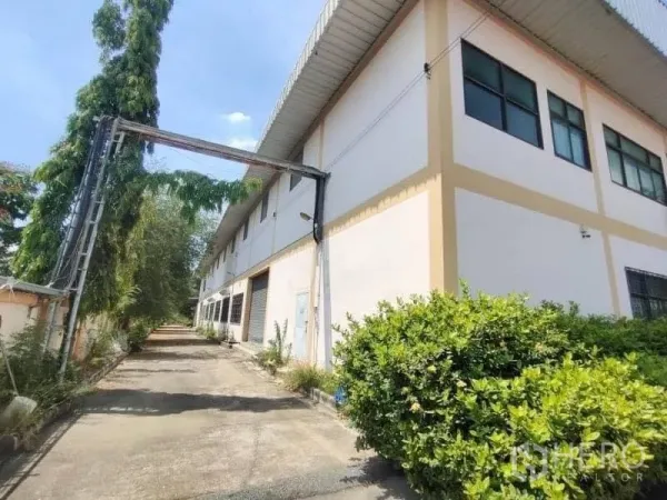 คลังสินค้า ขายในKhlong Toei, Bangkok - มุมด้านข้างอาคารโกดังสองชั้นหลังคาเมทัลชีท มีทางสัญจรและต้นไม้ ในวันที่ท้องฟ้าโปร่ง