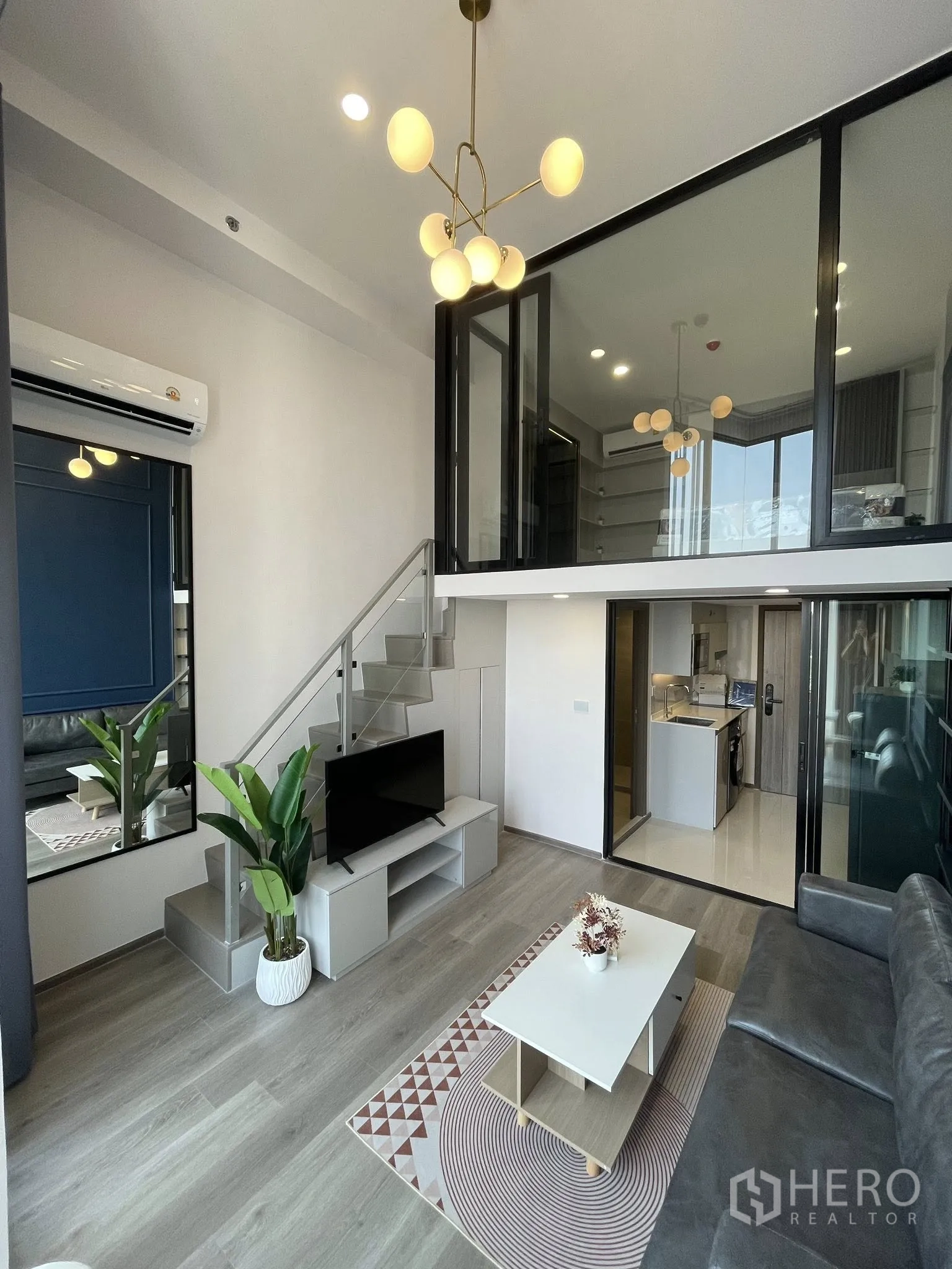 Condominium for rent in Khlong Toei, Bangkok - Duplex living space showing staircase to mezzanine, TV unit, coffee table, plant, and compact kitchen.