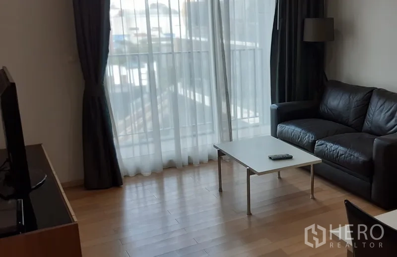 คอนโด ให้เช่าในKhlong Toei, Bangkok - ห้องนั่งเล่นคอนโดสว่าง โปร่ง เพดานสูง มีผ้าม่านทึบและโซฟาหนังสีดำข้างหน้าต่างระเบียง