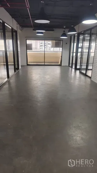 办公空间 出租在Pathum Wan, Bangkok - 开放式办公区，抛光水泥地、玻璃隔断与吊灯，落地窗通向阳台可见城市景观。