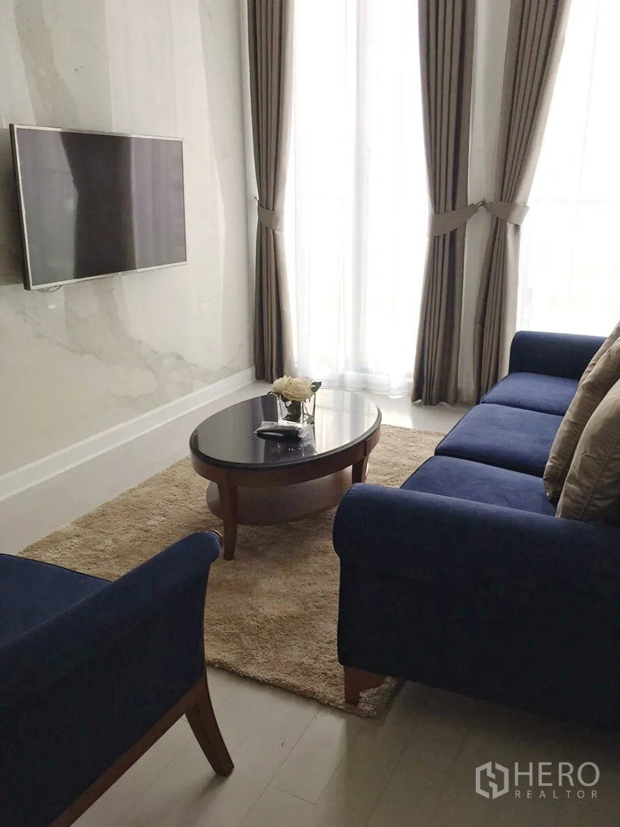 คอนโด ให้เช่าในPathum Wan, Bangkok - มุมนั่งเล่นอบอุ่น ทีวีติดผนัง โต๊ะกลางทรงวงรี และผ้าม่านสูงจรดเพดาน