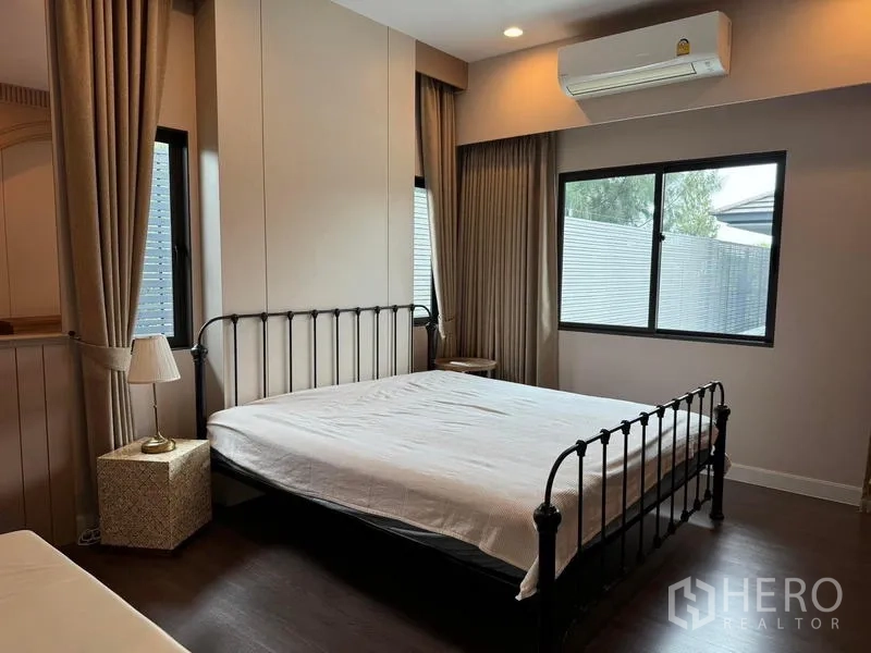 Detached House for rent in Bang Kapi, Bangkok - Cozy bedroom with metal bedframe, large windows, curtains and wall‑mounted air conditioner over wood flooring.