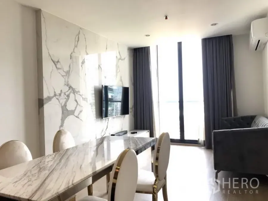 Condominium for rent or sale in Watthana, Bangkok - Bright living and dining area with marble accent wall, wall‑mounted TV, marble table, gray sofa, and floor‑to‑ceiling windows.