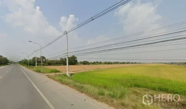 Land for sale in Pathum Thani - Wide roadside plot in Lam Luk Ka with flat green fields, power lines and streetlights under a clear sky.