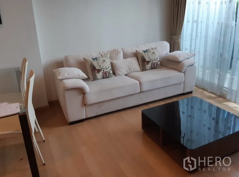 Condominium for rent in Khlong Toei, Bangkok - Bright living area featuring a white sofa, glass dining set, sheer curtains and sliding door to a balcony.