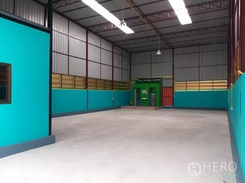 Warehouse for rent in Nonthaburi - Spacious warehouse hall with teal walls, bright lighting and a green restroom unit at the rear.