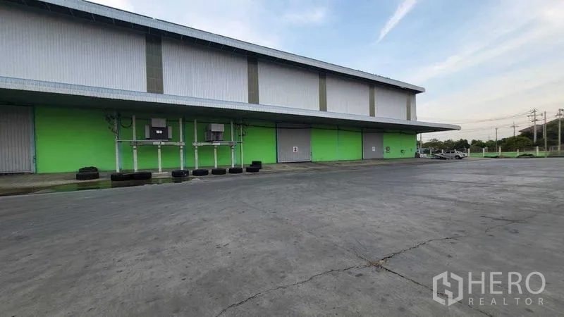 工厂 出租在Sai Noi, Nonthaburi - 侧立面为绿色墙体，设有多处卷帘门与管线设备，前方硬地宽阔。