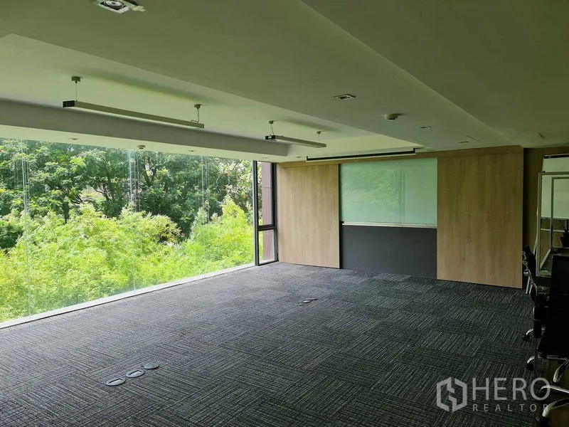 Office Space for rent in Watthana, Bangkok - Open-plan office with floor-to-ceiling windows, lush green views, carpet tiles, suspended linear lights and a wood panel wall with a writable board.