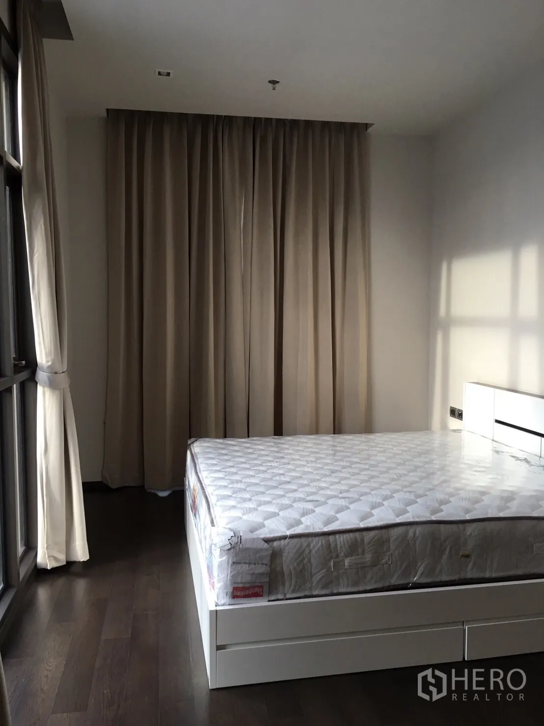 Condominium for rent in Watthana, Bangkok - Bright bedroom with white bed frame, dark wood floors, large windows and beige curtains.