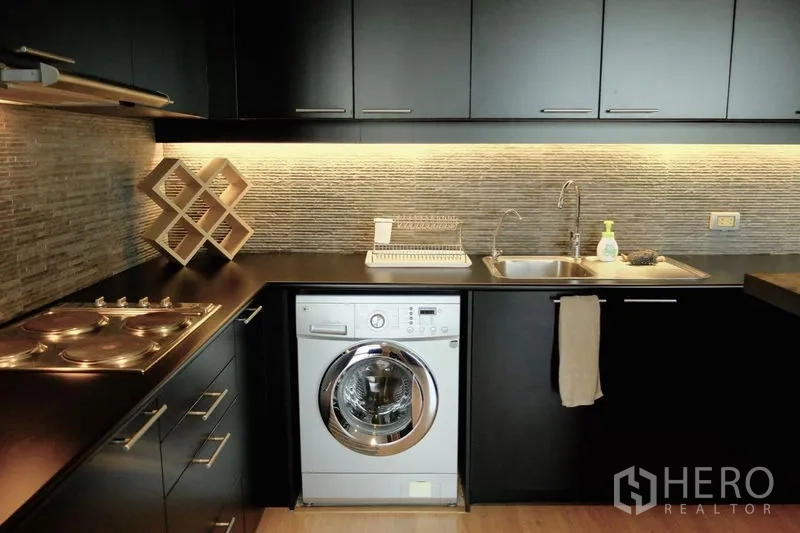 Condominium for sale in Watthana, Bangkok - Close-up of L-shaped kitchen with under-cabinet lighting, sink and a built-in washing machine.