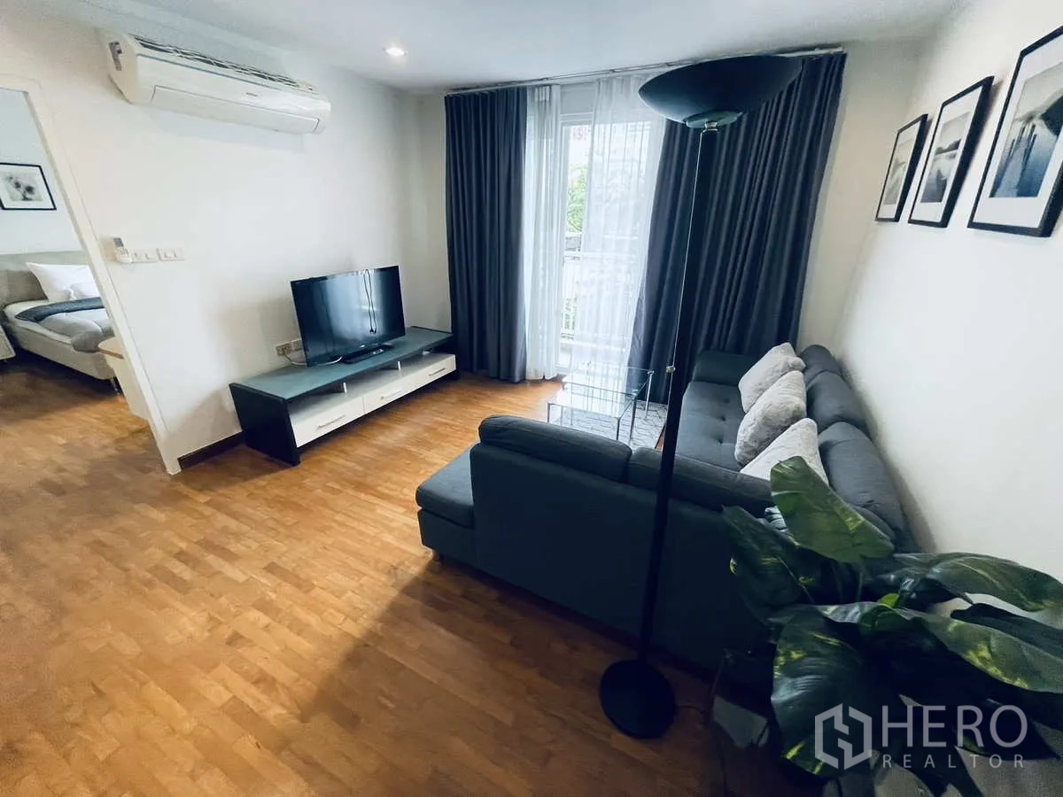 Condominium for rent in Watthana, Bangkok - Bright living room with gray sectional sofa, TV console, wood flooring, and balcony curtains.