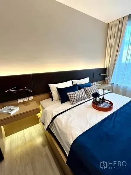 Condominium for sale in Bang Sue, Bangkok - Bright bedroom with blue-accent bedding, backlit headboard, bedside desk and large window.