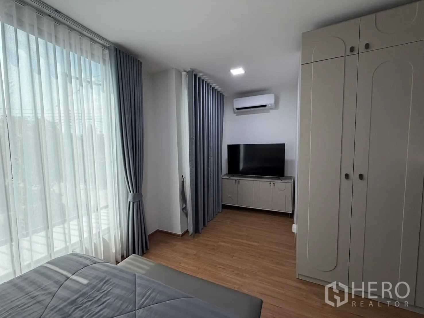 Townhouse for rent in Bang Phli, Samut Prakan - Primary bedroom corner with TV console, air-conditioner, wardrobes and long windows with grey curtains.
