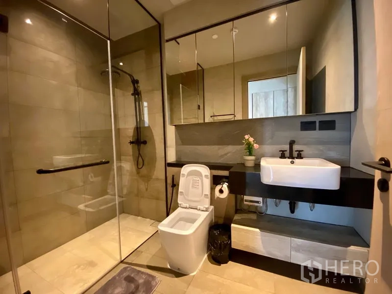 Condominium for rent in Watthana, Bangkok - Second bathroom with glass-enclosed shower, floating vanity, mirrored storage, and warm tiles.