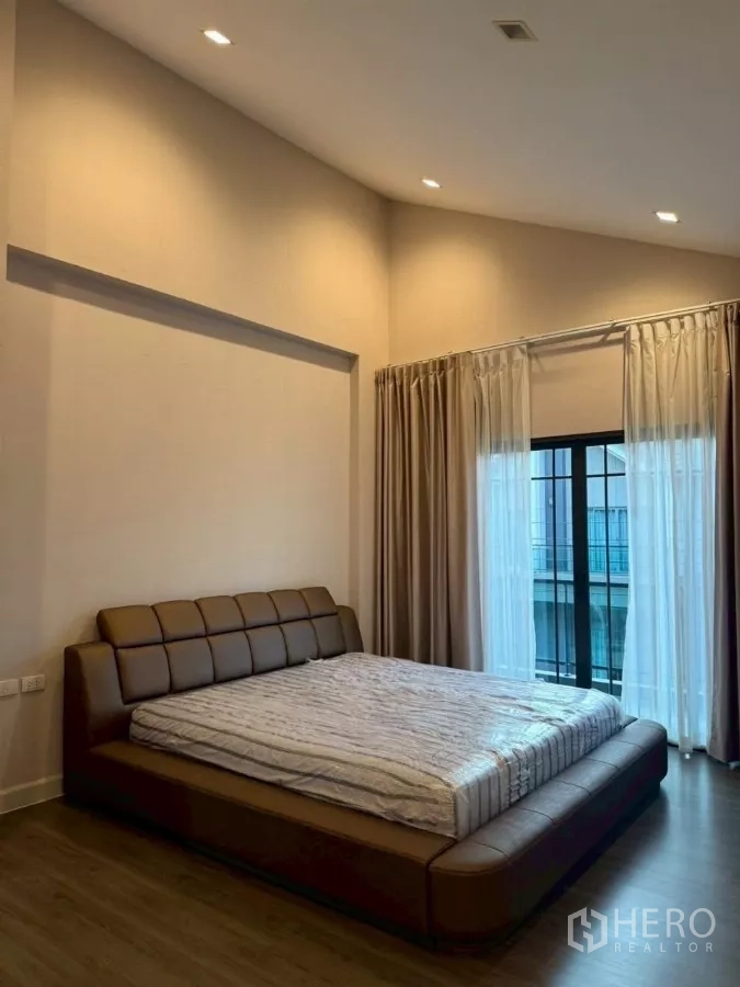 Detached House for rent or sale in Bang Phli, Samut Prakan - Master bedroom with high ceiling, large upholstered bed and sliding door to the balcony with sheer curtains.