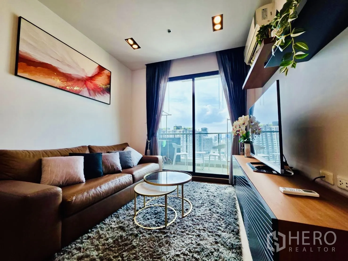 Condominium for rent in Huai Khwang, Bangkok - Cozy living room with leather sofa, nesting coffee tables, and sliding doors to a city-view balcony.