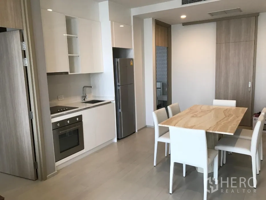 Condominium for rent or sale in Pathum Wan, Bangkok - Modern open-plan kitchen with white cabinetry, built-in oven, cooktop, sink, and a six-seat dining table.