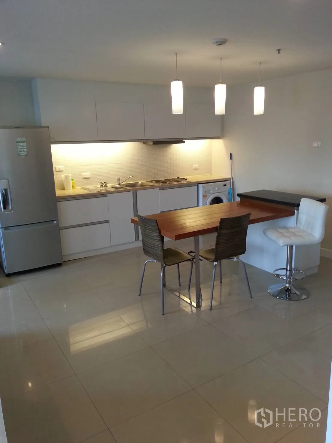 Condominium for rent in Ratchathewi, Bangkok - Open-plan kitchen with white cabinetry, pendant lights, dining table and washer.