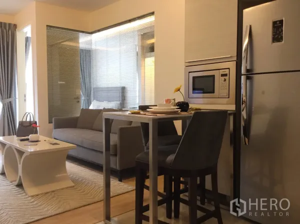 公寓 出租或出售在Watthana, Bangkok - 开放式起居区，灰色沙发与吧台用餐区，卧室由玻璃隔断分隔。