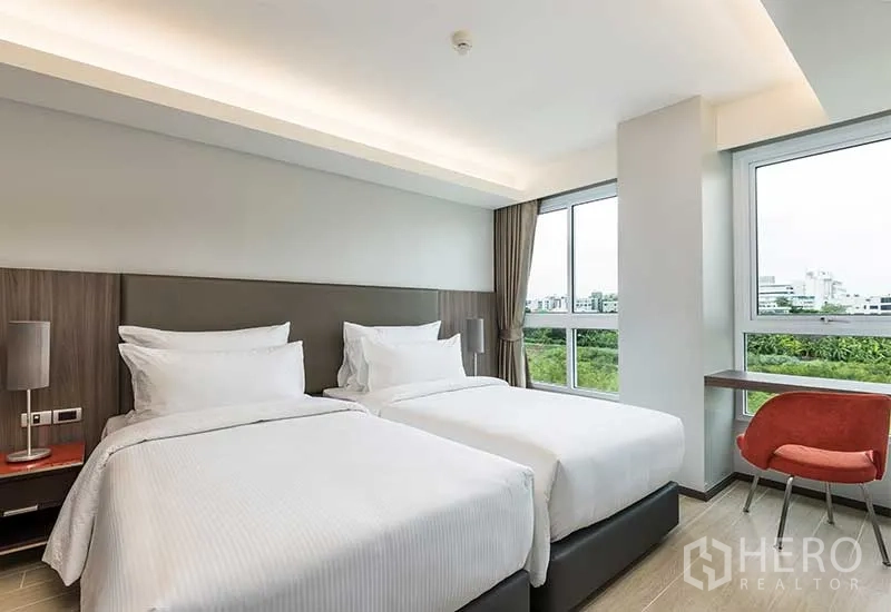Apartment for rent in Huai Khwang, Bangkok - Second bedroom with twin beds, study desk, and corner windows overlooking greenery and skyline.