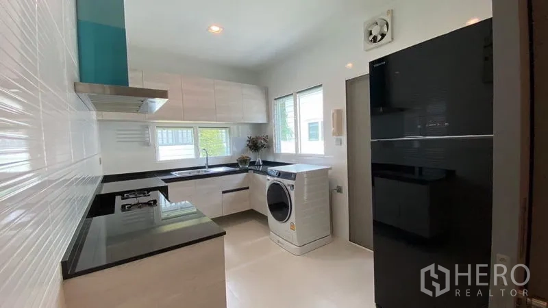 Detached House for rent in Samut Prakan - Fitted kitchen with black countertops, white cabinets, appliances and washing machine by windows.