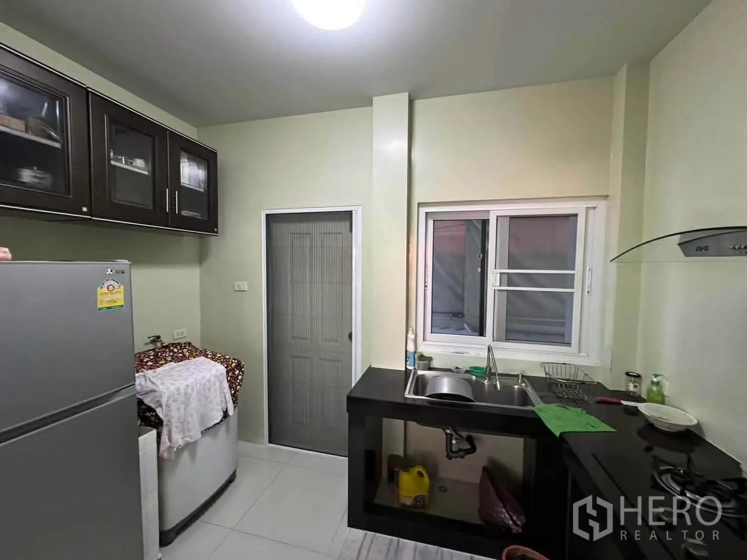 Detached House for rent in Watthana, Bangkok - Compact kitchen with sink, dark upper cabinets, gas cooktop, and a window.