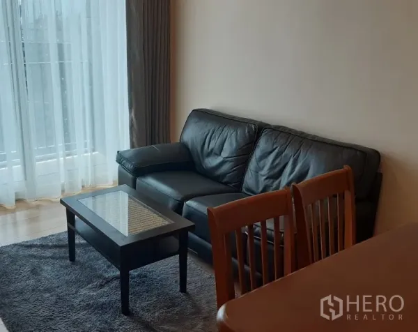 คอนโด ให้เช่าในKhlong Toei, Bangkok - มุมนั่งเล่นอบอุ่น โซฟาหนังสีดำ โต๊ะกลางกระจก ชุดโต๊ะทานอาหาร และหน้าต่างเต็มผนังพร้อมผ้าม่านโปร่ง