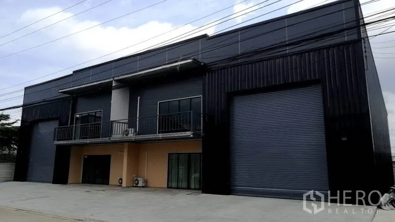 Warehouse for rent or sale in Lam Luk Ka, Pathum Thani - Modern black metal warehouse with two large roller shutters, balconies, and a wide parking apron in Lam Luk Ka, Pathum Thani.