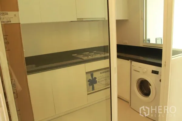 Condominium for rent or sale in Watthana, Bangkok - Enclosed kitchen with white cabinetry, black countertop, electric cooktop, and washing machine behind sliding glass doors.