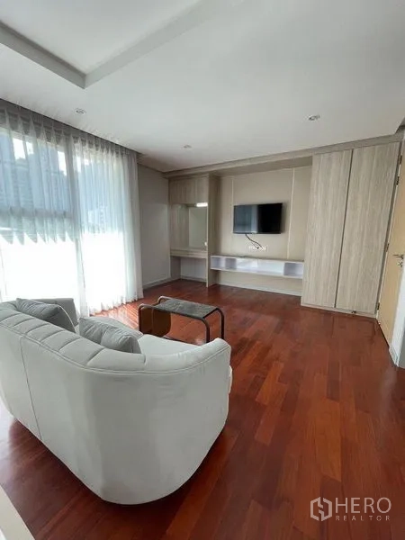 Condominium for rent in Watthana, Bangkok - Living room with light-colored sofa, built-in TV wall and large sliding windows with sheer drapes and wood floors.