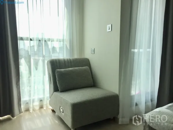Condominium for rent in Ratchathewi, Bangkok - Cozy grey lounge chair beside floor-to-ceiling windows with sheer curtains and city light filtering in.
