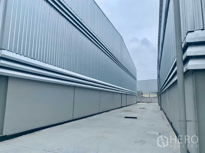 Warehouse for rent in Ban Bueng, Chon Buri - Service lane between warehouse buildings showing drainage openings and metal cladding.