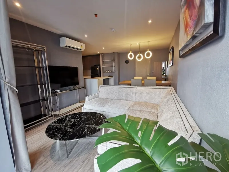 Condominium for rent or sale in Watthana, Bangkok - Modern open-plan living and dining room with an L-shaped sofa, black marble coffee table, wall-mounted TV, and circular pendant lights.